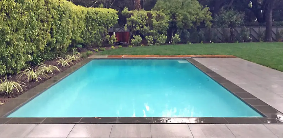 Remodeled Rectangular Swimming Pool With Bright Blue Water, Dark Tile Border, And Gray Concrete Deck Surrounded By Green
