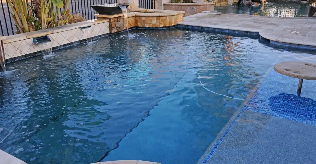 Remodeled Swimming Pool With Blue Tile Waterline, Stone Coping, Spa, And Water Features Surrounded By Pavers